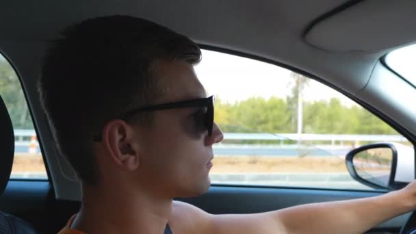 Een knappe vent met een zonnebril die 's zomers door de weg rijdt. Profiel van de mens zorgvuldig op zoek naar het verkeer en de controle van zijn voertuig. Concept van een road trip. Uitzicht van binnenuit auto - Video