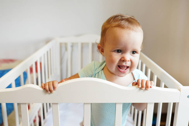 ベビーベッドに立っている美しい赤ん坊。可愛いです女の子の肖像画でコットと笑い - 写真・画像