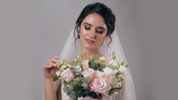 Jonge aantrekkelijke bruid in bruidsjurk met bruidssluier houdt boeket en raakt het op witte achtergrond. Meisje in trouwjurk. Advertentie voor sociale netwerken voor huwelijksbureau. - Video