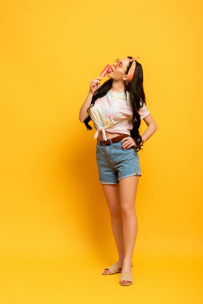 stylish summer brunette girl licking ice cream on yellow background - Photo, Image
