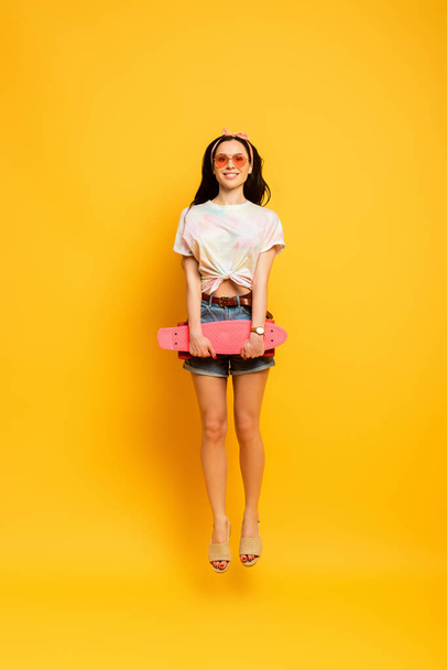 happy stylish summer brunette girl jumping with pink penny board on yellow background - Fotoğraf, Görsel