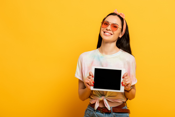 улыбается стильная летняя брюнетка с цифровым планшетом с чистым экраном на желтом фоне - Фото, изображение