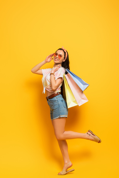 pełny widok długości uśmiechnięty stylowy lato brunetka dziewczyna z torby na zakupy na żółtym tle - Zdjęcie, obraz