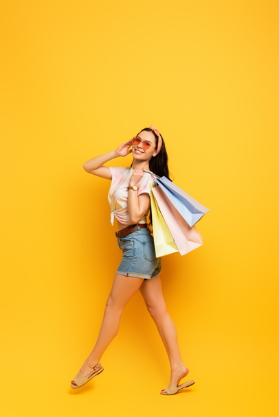 full length view of smiling stylish summer brunette girl walking with shopping bags on yellow background - Foto, Imagem