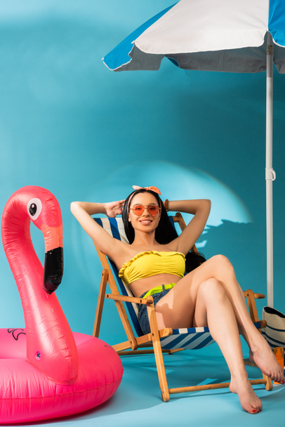 smiling stylish girl sitting in deck chair near inflatable flamingo, beach bag and umbrella on blue background - Fotó, kép