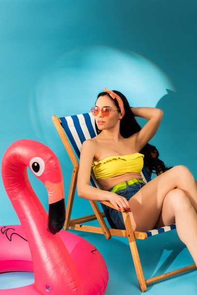 stílusos lány ül fedélzeti szék közelében felfújható flamingo kék háttér - Fotó, kép
