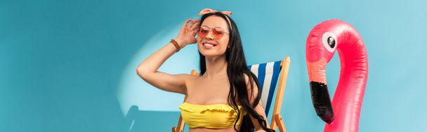 smiling stylish girl sitting in deck chair near inflatable flamingo on blue background, panoramic shot - Photo, Image