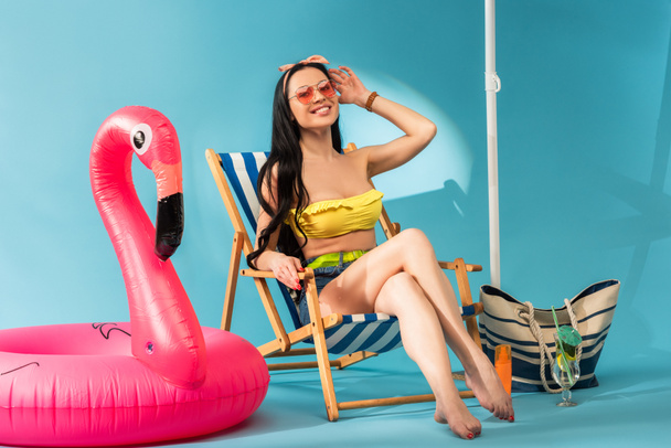 sonriente chica elegante sentada en la silla de cubierta cerca de flamenco inflable, bolso de playa y cóctel sobre fondo azul
 - Foto, Imagen