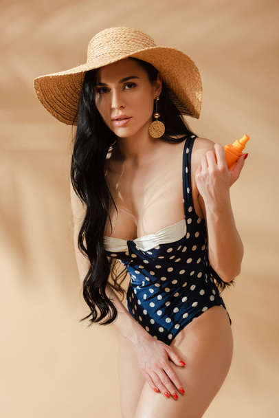 sexy femme brune en maillot de bain à pois et chapeau de paille appliquant de la crème solaire sur fond beige - Photo, image