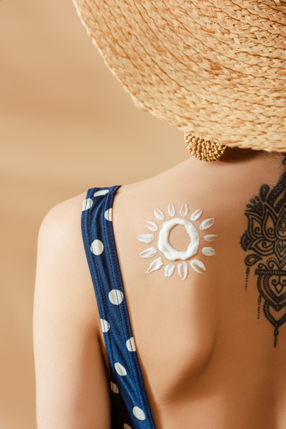 женщина в купальнике в горошек и соломенной шляпе с нарисованным солнцем и татуировкой на бежевом фоне - Фото, изображение
