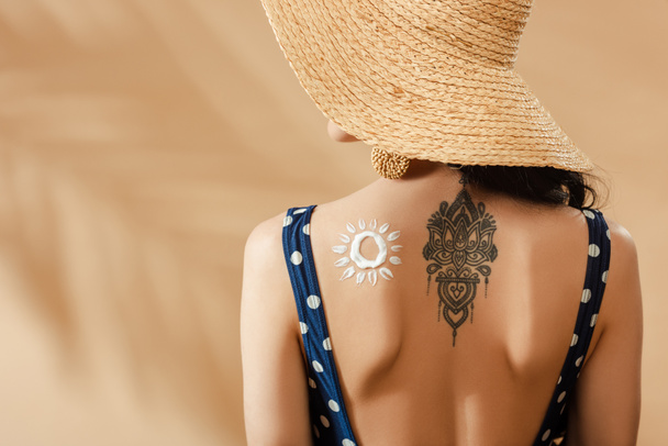woman in polka dot swimsuit and straw hat with drawn sun and tattoo on back on beige background - Photo, Image