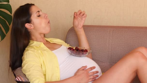 Young pregnant woman eating cherries sitting on the sofa. Beautiful pregnant girl eating cherries from a glass plate. - Materiał filmowy, wideo