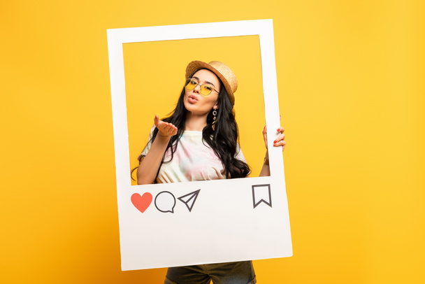 brunette meisje in de zomer outfit poseren blazen kus in sociaal netwerk frame op gele achtergrond - Foto, afbeelding