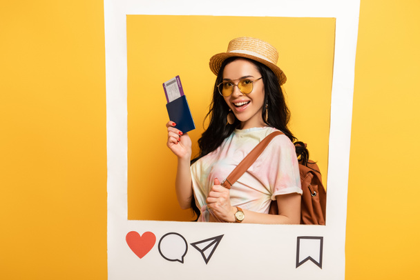 gelukkig brunette meisje in de zomer outfit houden vliegticket in sociaal netwerk frame op gele achtergrond - Foto, afbeelding