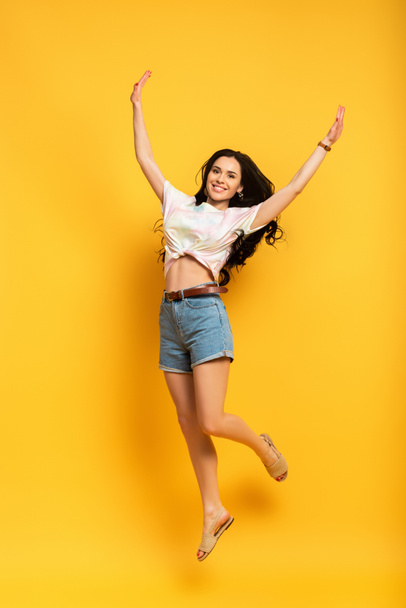 full length view of happy brunette girl jumping on yellow background - Foto, Bild