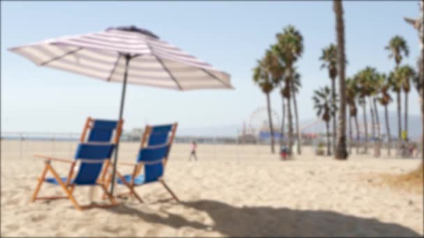 Spiaggia della California sfocata, due sedie a sdraio blu vuote, ombrellone a strisce vicino al molo di Santa Monica Pacific Ocean Resort. Summertime relax lounge atmosfera da sogno e palme, Los Angeles CA USA - Filmati, video