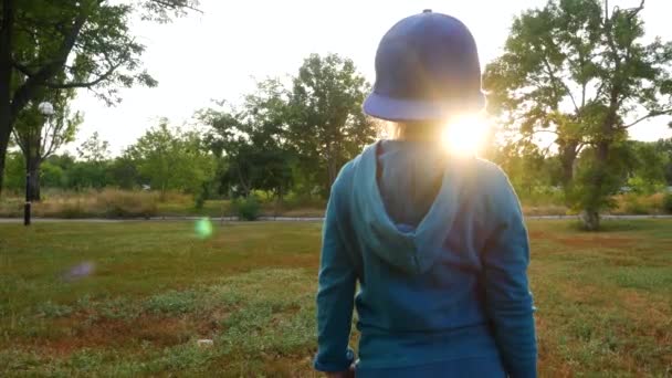 Cute child in blue hoody and snapback staying in autumn forest. Young girl walks on green grass rare back view faceless. Little kid walking on forest path on fresh air sunrays outdoor, sun light flash - Filmati, video
