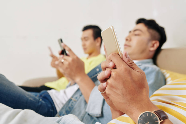Amici maschi asiatici che riposano sul divano a casa, messaggiano, leggono articoli e giocano su smartphone - Foto, immagini