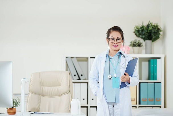 Portret van een zelfverzekerde vrouwelijke arts die klembord vasthoudt wanneer hij aan haar bureau staat - Foto, afbeelding