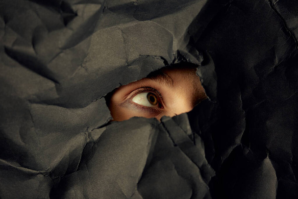 Womans eye in hole in black crumpled paper background, minimalist concept photography for blog or poster - Photo, Image