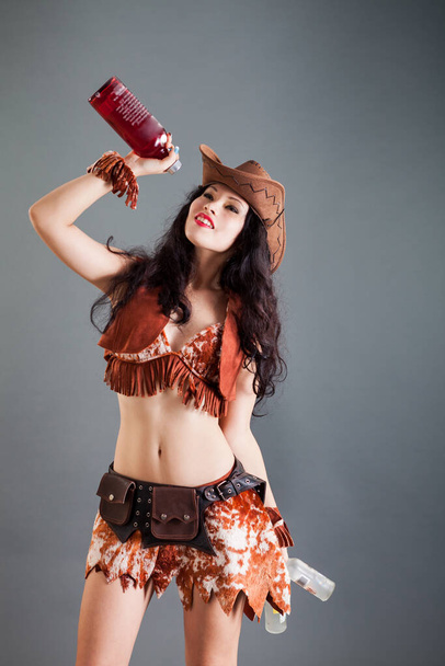 jolie fille brune en costume de cow-boy franc et pose de chapeau à la caméra avec des bouteilles dans les mains sur fond gris en studio - Photo, image