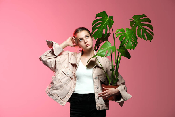 A girl in a pink jacket in black jeans holds in her hands a monstera flower in a pot posing on a pink background. - Foto, Imagen