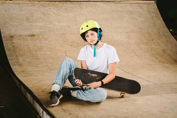 Schoolgirl after lessons at skateboarding practice in outdoor skate park. Stylish and beautiful caucasian girl with skateboard on a half pipe ramp in a skatepark. Teenager skater girl with skate deck. - Photo, Image