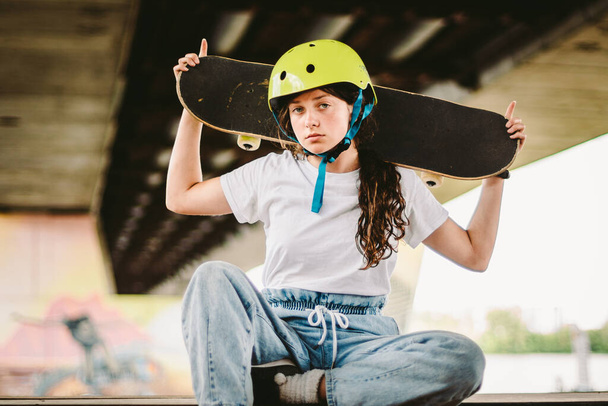 Έφηβη κοπέλα με κράνος και κομψά ρούχα που ποζάρουν σε μισή ράμπα σωλήνα ένα υπαίθριο πάρκο για σκέιτ. Όμορφο παιδί θηλυκό μοντέλο skateboarder με πατινάζ του σκάφους στο αστικό ακραίο πάρκο. Μαθήτρια μετά το σχολείο.. - Φωτογραφία, εικόνα