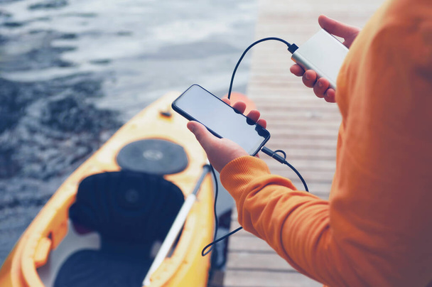 The guy holds a portable charger with a smartphone in his hand. A man against the background of water and a kayak charges the telephone with Power Bank. Concept on the theme of tourism - Photo, Image