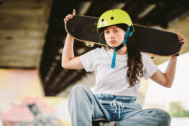Schoolgirl after lessons at skateboarding practice in outdoor skate park. Stylish and beautiful caucasian girl with skateboard on a half pipe ramp in a skatepark. Teenager skater girl with skate deck. - Photo, Image