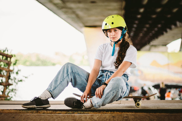 Portrait confident, cool young female skateboarder at outdoor skate park. Urban girl with skate board on half pipe ramp. lifestyle. Teen model skateboarder posing in helmet and stylish clothes. - Фото, изображение