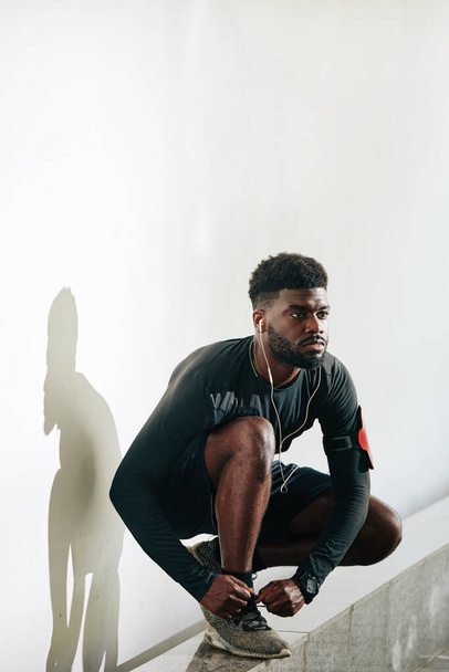 Seriöser junger schwarzer Sportler bindet Schnürsenkel, wenn er sich auf den Sprint-Triathlon vorbereitet - Foto, Bild