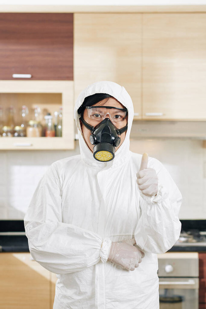 Portret van een professionele schoonmaakster in een beschermend hazmatpak in een gedesinfecteerde keuken met duimen omhoog - Foto, afbeelding