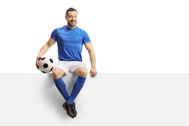 Footballer in a blue jersey with a soccer ball sitting on a blank panel and smiling isolated on white background - 写真・画像