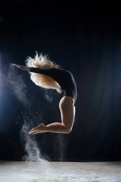 Beautiful plump blonde girl wearing a black gymnastic bodysuit covered with clouds of the flying white powder jumps dancing sitting on a dark. Artistic conceptual and advertising photo. Copy space - Foto, Imagem