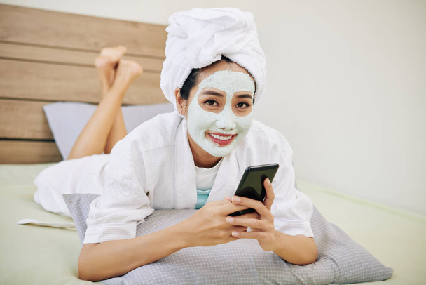 Feliz joven asiática mujer con máscara de arcilla en su cara descansando en la cama y mensajes de texto amigos
 - Foto, Imagen