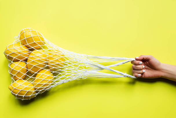 cropped view of woman holding ripe whole lemons in string bag on yellow background - Foto, imagen