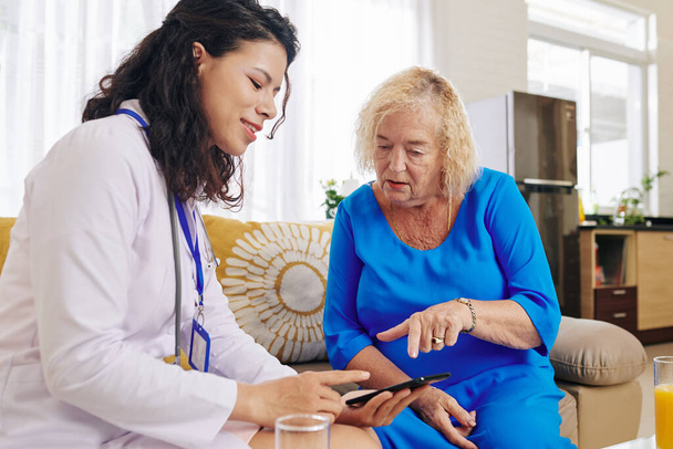 Médecin souriant visitant la patiente âgée à la maison et lui expliquant comment utiliser l'application de soins de santé pour communiquer avec le médecin - Photo, image