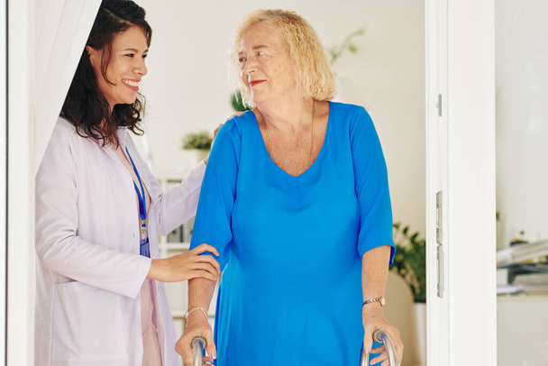 Usmívající se hezká zdravotní sestra pomáhá její starší pacient používat přední-kolové chodítko - Fotografie, Obrázek