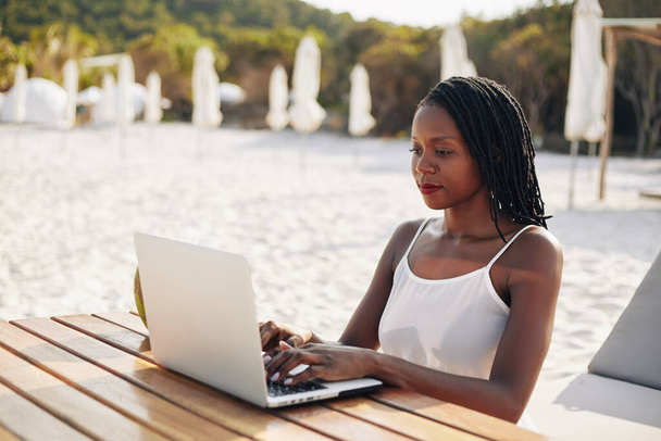 Sorrindo muito jovem blogueiro de viagens sentado à mesa de madeira na praia e trabalhando em laptop ao fazer post para seu blog
 - Foto, Imagem