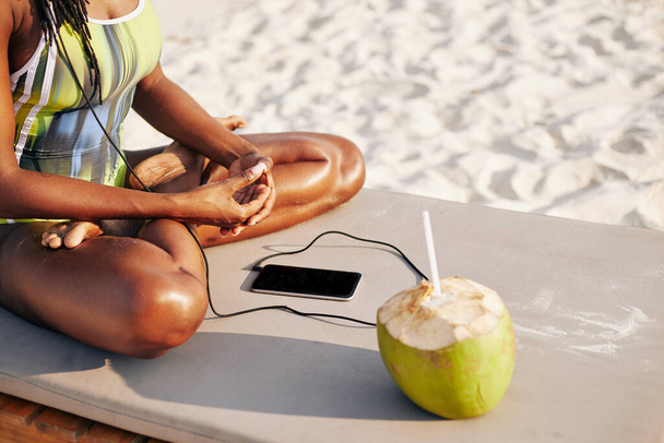 Cropped image of young fit Black woman sitting on chaise-lounge, drinking coconut water and listening to music in her smartphone - Photo, Image