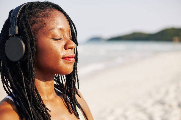 Лицо красивой молодой чернокожей женщины, улыбающейся и закрывающей глаза, слушая расслабляющую музыку в наушниках и наслаждаясь морским бризом - Фото, изображение