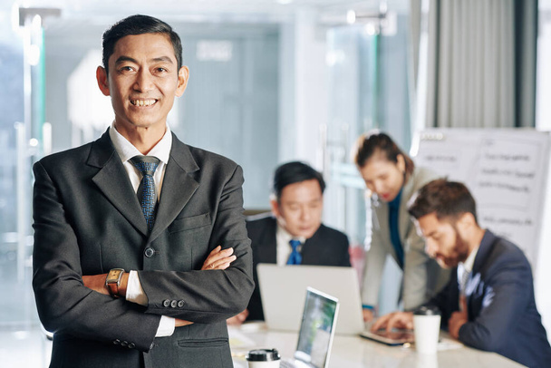 Porträt eines lächelnden selbstbewussten asiatischen Unternehmers, der mit verschränkten Armen dasteht, während seine Kollegen im Hintergrund an der Strategie zur Geschäftsentwicklung arbeiten - Foto, Bild