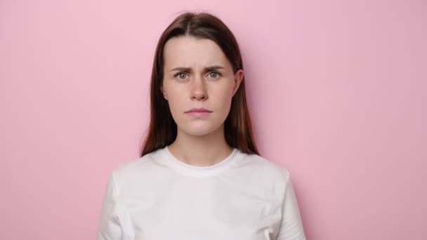 Displeased young annoyed woman pointing at camera and showing stupid gesture, blaming some idiot, dressed in white t-shirt, isolated on pink studio background. Brunette girl in despair and shock - Кадри, відео