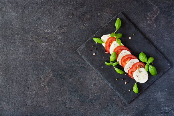 Caprese salát s mozzarelou, rajčaty, čerstvým olivovým olejem a bazalkou na břidlicové desce na černém pozadí horní pohled. Lahodný italský veganský salát. - Fotografie, Obrázek