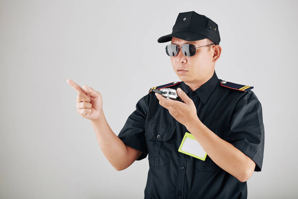 Poważny policjant w okularach przeciwsłonecznych za pomocą krótkofalówki lub radia policyjnego podczas demonstracji i kontroli pracy oficerów - Zdjęcie, obraz