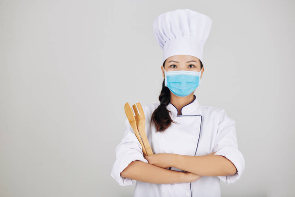 Σίγουρος νεαρός σεφ εστιατορίου με ιατρική μάσκα ποζάρει με ξύλινα σκεύη - Φωτογραφία, εικόνα