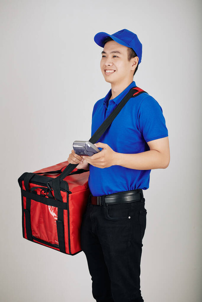 Portrait of handsome happy young courier carrying payment terminal when delivering food - Photo, Image