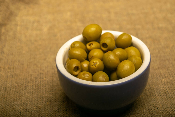 Green olives in a ceramic bowl on a background of coarse-textured burlap. Close up - Photo, Image