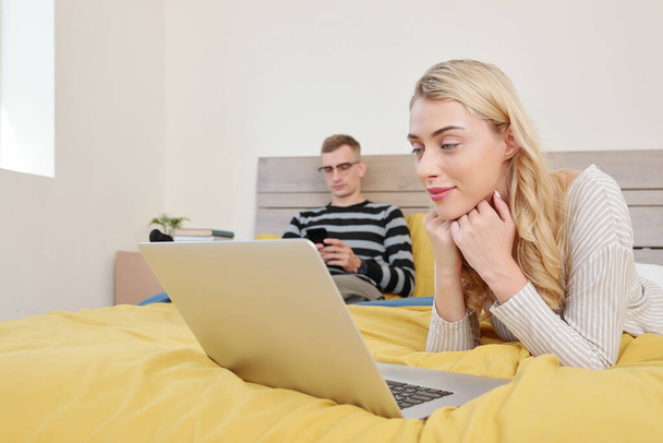 Ładna młoda uśmiechnięta kobieta odpoczywająca w łóżku i oglądająca nowy odcinek serialu na laptopie, gdy mąż pisze do przyjaciół - Zdjęcie, obraz